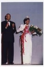 A young woman, wearing a floor-length dress with a tiara and bouquet of flowers, stands with Dr. Thomas W. Cole, Jr. on stage.