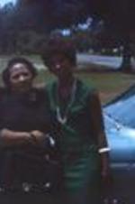 Two unidentified women pose in front of a car in a driveway. Unknown location.