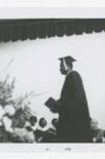 A man, wearing a graduation cap and gown, walks across the stage at commencement.