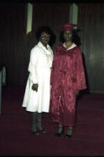 An unidentified young woman in a red cap &amp; gown stands next to an older woman, possibly her grandmother, on a small short staircase.