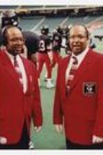 Pharrol Floyd and Darrol Floyd stand in front of football players on a football field.