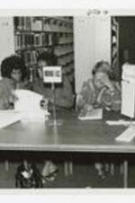Three women sit at a long table with books and signs "Home Ec." in a library.
