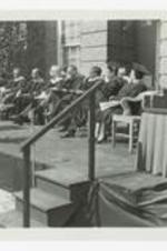 Men and women, wearing graduation caps and gowns, sit on an outdoor stage at commencement.
