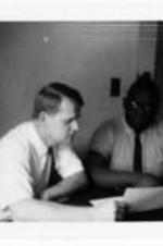 James Costen and an unidentified man sit at a table and read off a sheet of paper.
