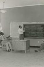 A teacher writes on a chalk board in a classroom of students.
