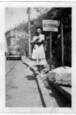An unidentified woman stands on the curb of a town street. She stands next to a sign reading: "Positively No Parking. 10.00 fine." A car is parallel parked behind her.