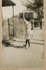 A girl carries a large basket on her head and walks through a garden gate. Written on recto: Girl at the open gate.