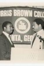 Two men at a podium, flag in background: Morris Brown College. Written on verso: "L to R; Head football coach- Gregory Thompson; Head Basketball Coach- Harold Merrill".