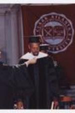 A man receives a graduation hood from a woman on stage at commencement.