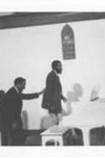 An unidentified man leads Dick Gregory around a piano at an event.
