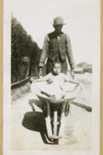 A young girl rides in a wheelbarrow pushed by a man.