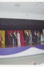 Young men and women, wearing formal wear with tiaras and sashes, stand in pairs on stage.