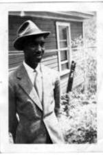 An unidentified man wearing a suit, tie, and fedora stands in front of a house.
