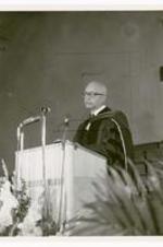 An unidentified man speaks at a podium wearing a commencement robe.