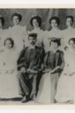Eleven women, wearing matching dresses, pose with a man and another woman, wearing graduation caps and gowns.