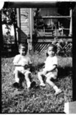 Two unidentified children sit in the grass of in front of an unidentified house.