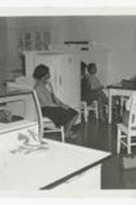 A woman sits at a table, while five other women look on from chairs, a poster hangs on the wall in the background in a home economics classroom.