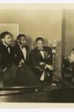 Five men, wearing suits with bow ties, pose around a grand piano with a microphone "CBS" in a room.