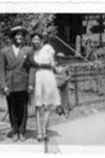 An unidentified couple stand on the sidewalk in front of an large house with an iron fence around the front yard. An unidentified boy is standing behind them, wearing a suit and a hat.
