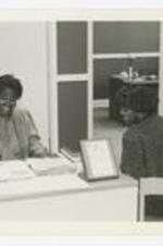 Two female staff members speak with each other at a desk.