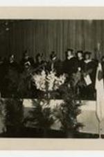 Men and women, wearing graduation caps and gowns, stand on stage at commencement.