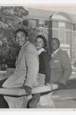 A young woman and two men sit on a bench outside of a brick building.
