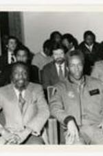 Dick Gregory with Guion Bluford seated in front of unidentified persons. Written on verso: Dick Gregory and Lt. Guion Bluford at Youth Motivational Rally in King Chapel -- January 13, 1984.