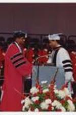 Four men stand on stage wearing red, white and black graduation cap and gowns.