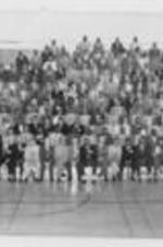 A group protrait of unidentified people sit on bleachers on a basketball court.