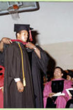An unidentified faculty member places a red hood on a graduate in front of platform participants.