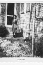 An unidentified woman stands in front of a brick house.