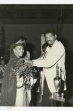 View of man and woman holding bouquet of flowers. Written on verso: "L to R Michelle Patmon, Dr. Colvert H. Smith; Dr. Smith ties Miss MBC cape- 1984-85"