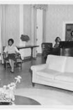 Two women sit in chairs while a man and a woman play at the piano. Written on verso: Students relaxing in University Dormitory.