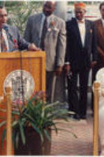 Southern Christian Leadership Conference (SCLC) President Joseph E. Lowery is shown speaking at an podium during an event outdoors that was part of the 35th Annual SCLC Convention. Coretta Scott King looks on and listens .Written on verso: SCLC/WOMEN