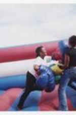 Two young women stand in an inflatable red, white and blue boxing ring bounce house with over-sized boxing gloves.