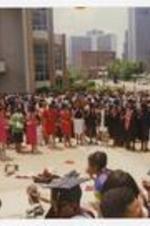 A large crowd of people stand in a circle outside, including graduates wearing graduation caps and gowns.