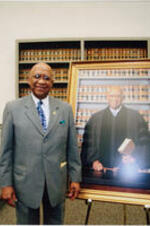 Judge John H. Ruffin, Jr. stands next to his portrait. Written on verso: Judge John H. (Jack) Ruffin, Jr. with portrait; A Tribute to the Honorable Judge John H. Ruffin, Jr. with Unveiling of Portrait, State Courts and Superior Court Courtroom Waynesboro, Georgia April 3, 2008