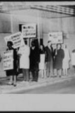 A group of protesters stand outside of a theatre on the sidewalk holding signs that read: "We're Willing to Die, But Not For Segregation", and "Negroes in Berlin? Yes, Negroes in the Dinkler? No".