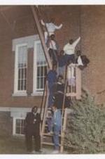 Outdoor group portrait of young men and women on fire-escape ladder. Written on verso: "1986 Brownite Yrbk Staff; From Top to bottom- Phillip Tate, Yvette Paris, Todd Blackburn, Travis Goodson, Shawnee Jackson, Michael Childress, John Jackson, Guinee Cook, Vincent E. Harris, Editor in Chief, Mr. Charles Barker, Consultant".