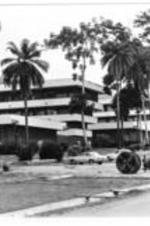 A boy stands next to an oversized cable spool that sits in a small parking lot. Behind them, a large building complex looms surrounded by tall Palm trees.