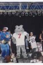 Two men hold a microphones on stage, a disc jockey, a grey dog mascot and other men stand in the background.