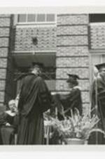Two men and a woman, wearing graduation caps and gowns, stand on an outdoor stage at commencement.