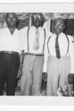 Three unidentified men stand together in a living room.  The man in the middle has his arms around the other men.