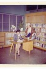 Ann Chandler sitting at desk next to unidentified woman in library. Written on verso: Ann autographing newly released index - Hunter St. Library.