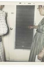 Two unidentified women stand in front of a door. One woman has curlers in her hair.