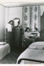 Interior of a women's dormitory room with one woman standing and another sitting at a desk. Written on verso: Atlanta University Dormitory scene, 1950