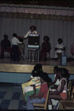 An unidentified woman stands on stage in front of children. The children sit in rows of wooden chair in front of the stage. Other unidentified adult sit in folding chairs behind the unidentified woman. The unidentified woman is handing an unidentified boy an unidentified object.
