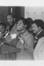 Maggie Bozeman is shown addressing a crowd alongside Julia Wilder (on left), Johnnie Carr (on right), and others in Tuskegee, Alabama. Written on verso: Mrs. Maggie Bozeman is joined by Mrs. Wilder, Rev. John L. Alford, Alabama Rep. Alvin Holmes, SCLC Board member Tuskegee Mayor Johnnie Ford, and SCLC Board member Mrs. Johnnie R. Carr, as the "homecoming" celebration begins in Tuskegee.