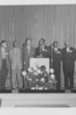An unidentified group of men stand on a stage.