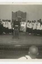 A man stands at a podium in front of a choir holding candles on a stage.